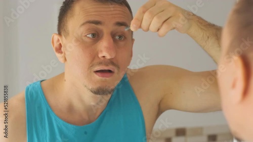 Middle-aged man plucks excess hair on eyebrows with tweezers and carefully examining himself in mirror. View over the shoulder.