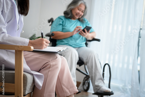 The doctor writing report for record the patient s condition  with blur background of Asian female elderly patient sitting in a wheelchair and using a mobile phone  to elderly and health care concept.