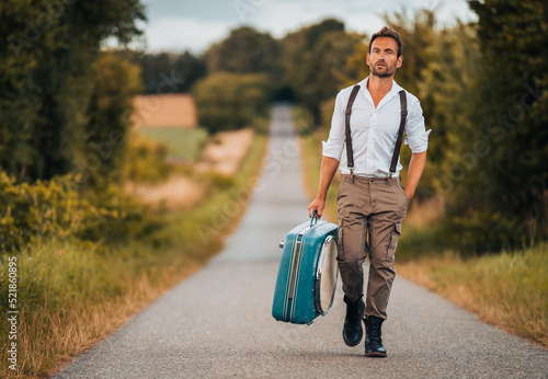 Individual man in vintage clothing holding a suitecase is walking along a street in nature photo