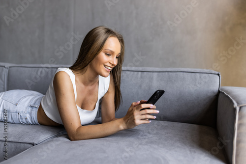 Smiling young woman using mobile phone while sitting on a couch at home with laptop