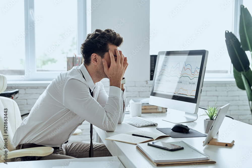 Depressed young man covering face with hands while sitting at his working place in office Depressed young man covering face with hands while sitting at his working place in office