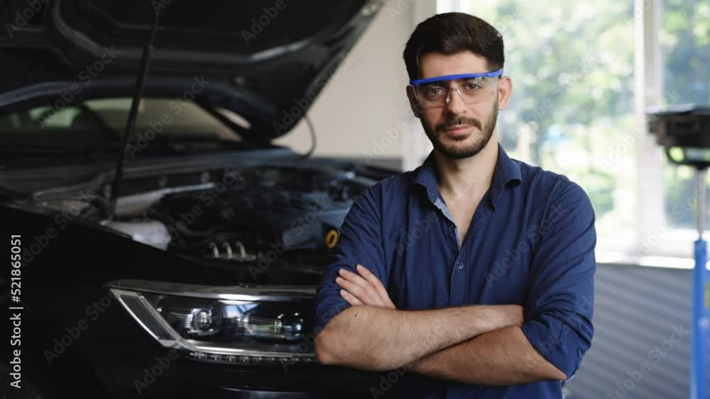 Portrait Bearded Handsome Car Mechanic is Posing in a Car Service. He Wears a Jeans Shirt and Safety Glasses. His Arms are Crossed. Specialist Manager Looks at a Camera and Smiles Portrait Bearded Handsome Car Mechanic is Posing in a Car Service. He Wears a Jeans Shirt and Safety Glasses. His Arms are Crossed. Specialist Manager Looks at a Camera and Smiles