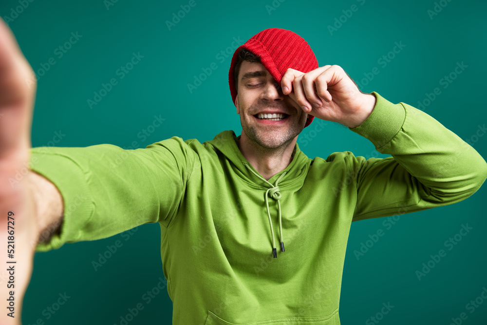 Fototapeta premium Happy young man making selfie and pulling hat on face while standing against green background