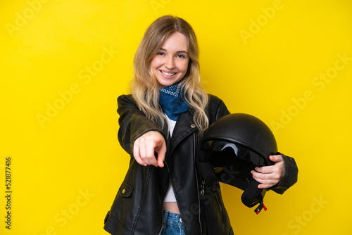 Blonde English young girl with a motorcycle helmet isolated on yellow background pointing front with happy expression