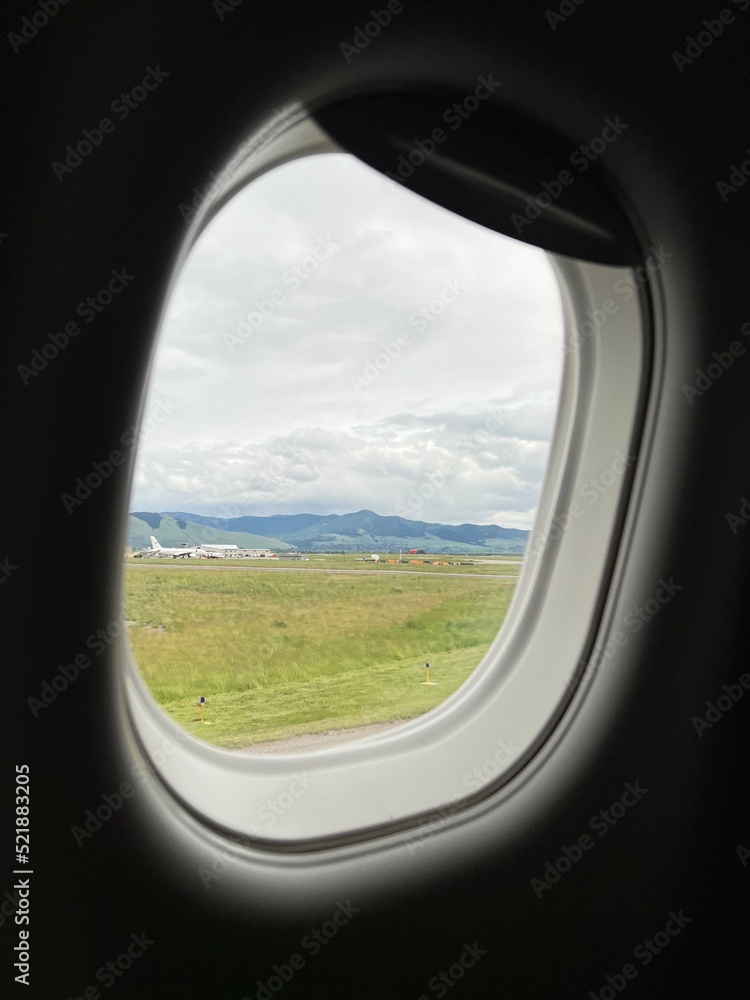 Mountains, clouds, and a distant aircraft seen through passenger jet window while taxiing to runway at Missoula Montana Airport