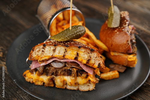 Delicious beef burger cut in half with cheese, pickled, tomatoes, mustard, and fries on a black plate on wooden background. Fast food.