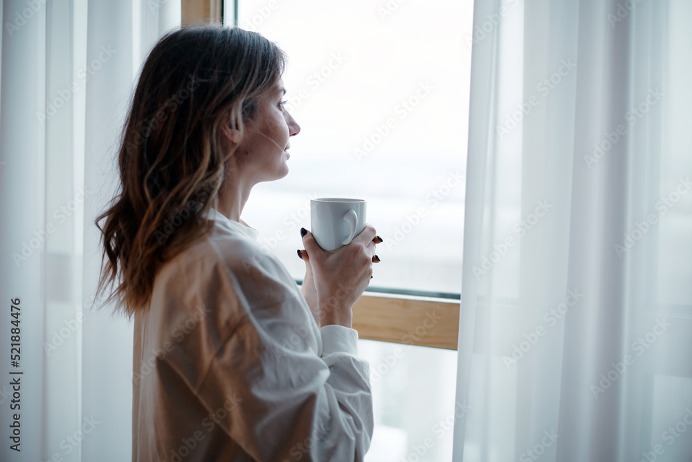 Romantic dreamy female homeowner tenant holding mug of hot coffee looking out of window. Leisurely morning Romantic dreamy female homeowner tenant holding mug of hot coffee looking out of window. Leisurely morning