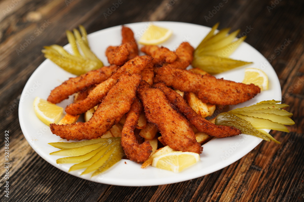 Crispy deep-fried chicken strips. Fried chicken strips on fried potatoes with pickles. Fried chicken and potatoes on a white plate on wooden background. Crispy deep-fried chicken strips. Fried chicken strips on fried potatoes with pickles. Fried chicken and potatoes on a white plate on wooden background.