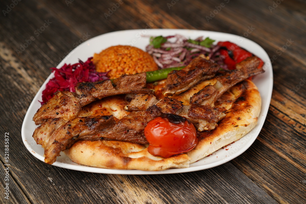 Grilled lamb chops on a white plate on wooden background. Grilled lamb with pita, garnish, onions, bulgur, roasted green pepper, and roasted tomato. Turkish name: kuzu pirzola. Grilled lamb chops on a white plate on wooden background. Grilled lamb with pita, garnish, onions, bulgur, roasted green pepper, and roasted tomato. Turkish name: kuzu pirzola.