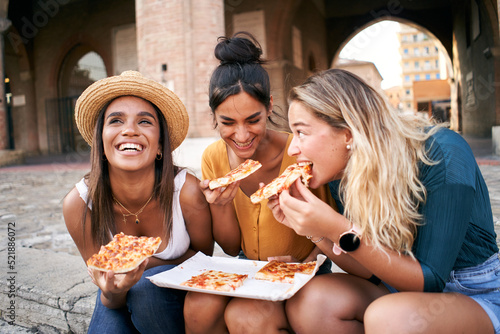 Funny mixed race group of three women eating street food outdoors in the city street. Happy female friends enjoying the summer vacations together European city. Young people lifestyle concept