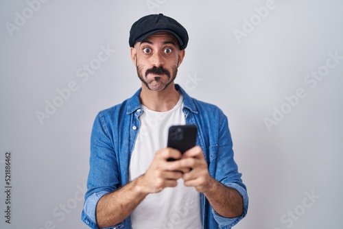 Hispanic man with beard using smartphone typing message puffing cheeks with funny face. mouth inflated with air, catching air. © Krakenimages.com