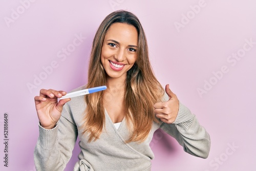 Beautiful hispanic woman holding pregnancy test result smiling happy and positive  thumb up doing excellent and approval sign