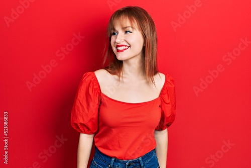 Redhead young woman wearing casual red t shirt looking away to side with smile on face, natural expression. laughing confident.
