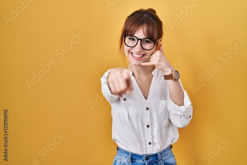 Bild auf Leinwand Young beautiful woman wearing casual shirt over yellow background smiling doing talking on the telephone gesture and pointing to you