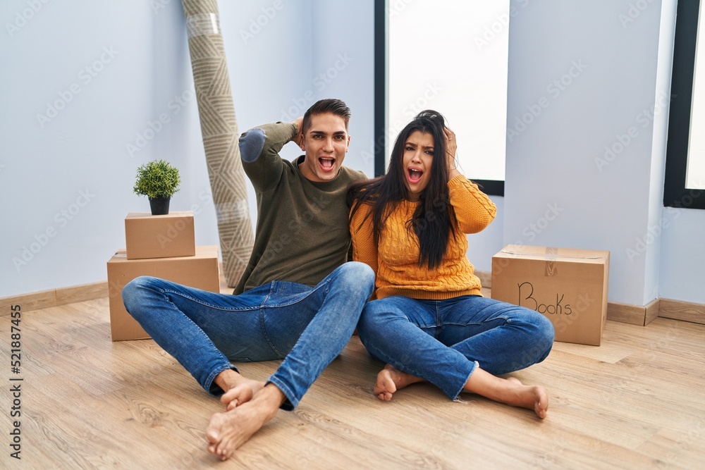 Young couple sitting on the floor at new home crazy and scared with hands on head, afraid and surprised of shock with open mouth