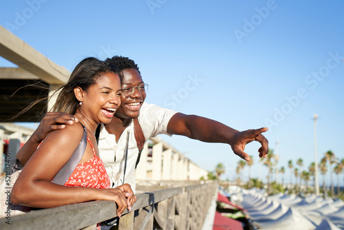 Beautiful couple of African origin have fun embracing in a coastal city center, leaning out of a gazebo pointing to the horizon. Background with copy space.