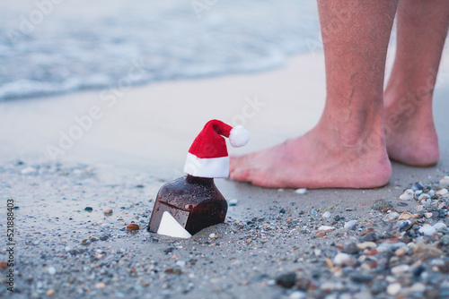 Close up view. Seasonal holidays concept. Single bottle of whisky with Santa hat in sand of the sea, Christmas party, Xmas time, festive decoration, soft focus. Seasonal holidays concept.
