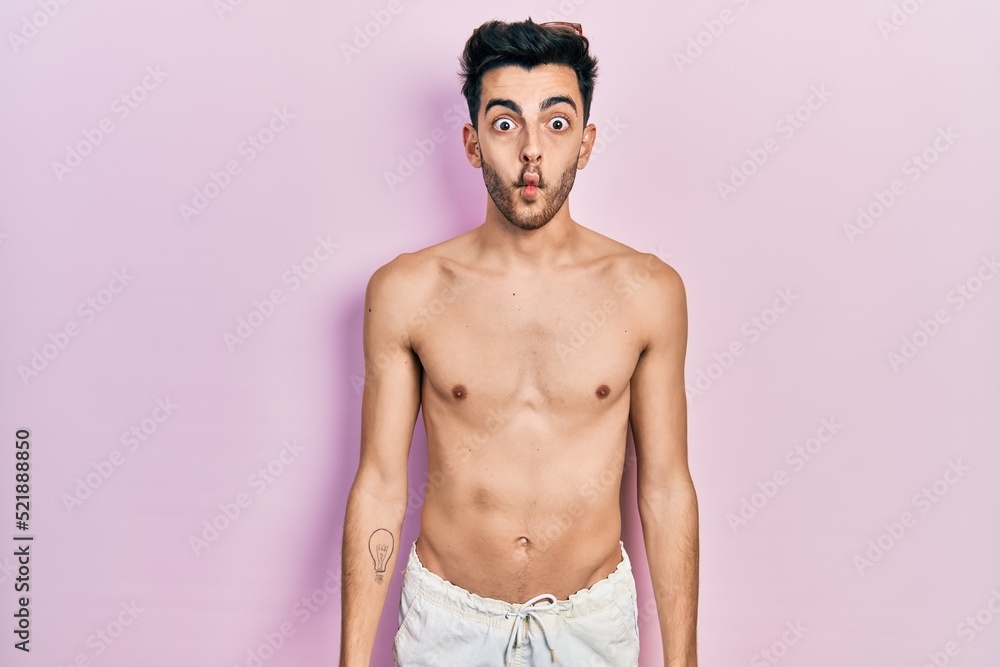 Fototapeta premium Young hispanic man wearing swimwear shirtless making fish face with lips, crazy and comical gesture. funny expression.