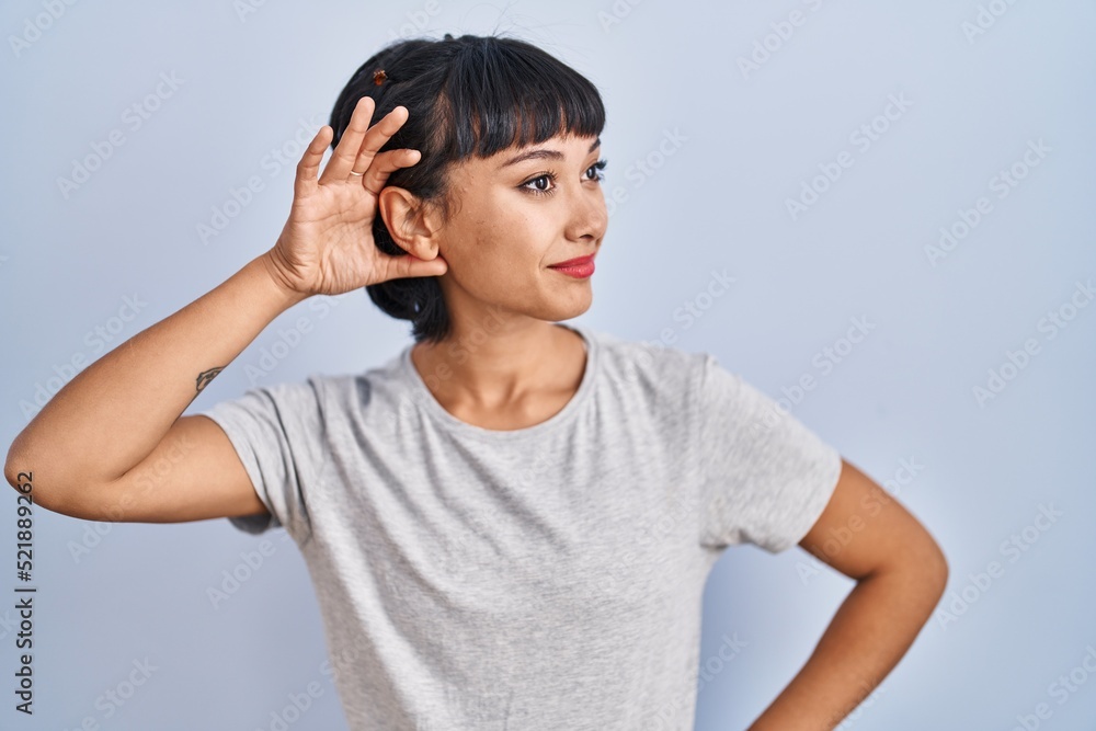Young hispanic woman wearing casual t shirt over blue background smiling with hand over ear listening an hearing to rumor or gossip. deafness concept. Young hispanic woman wearing casual t shirt over blue background smiling with hand over ear listening an hearing to rumor or gossip. deafness concept.