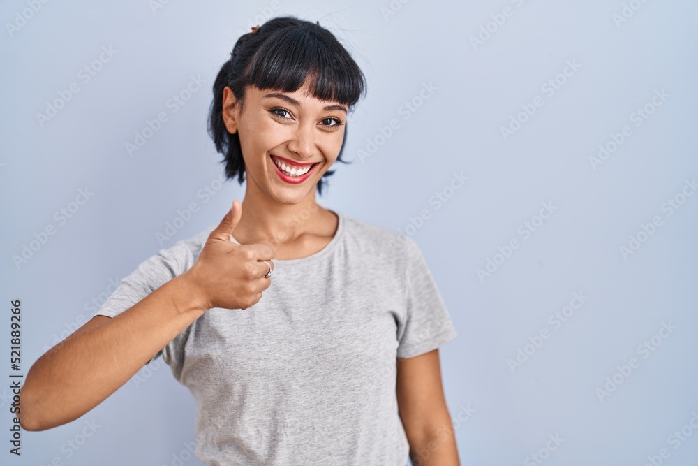 Young hispanic woman wearing casual t shirt over blue background doing happy thumbs up gesture with hand. approving expression looking at the camera showing success. Young hispanic woman wearing casual t shirt over blue background doing happy thumbs up gesture with hand. approving expression looking at the camera showing success.