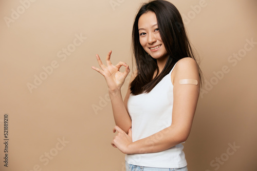 Enjoyed cute tanned adorable young Asian lady after vaccination show Okay gesture posing isolated on beige pastel background. People Emotions. CoVID Vaccination concept. Copy Space Offer Banner.