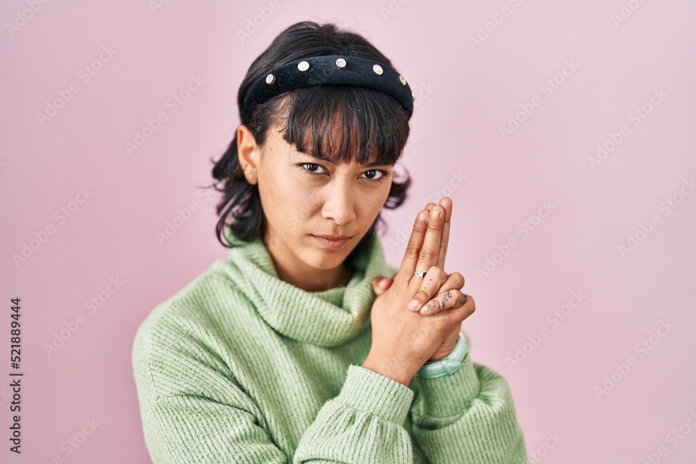 Young beautiful woman standing over pink background holding symbolic gun with hand gesture, playing killing shooting weapons, angry face Young beautiful woman standing over pink background holding symbolic gun with hand gesture, playing killing shooting weapons, angry face