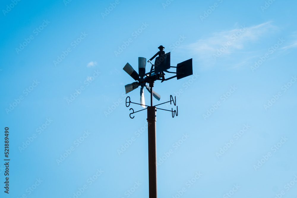 Anemometer, instrument to measures wind speed and direction. Fun weather station on a pole against blue sky with spinning wheel and cardinal directions. Construction includes cute cyclist figure. Anemometer, instrument to measures wind speed and direction. Fun weather station on a pole against blue sky with spinning wheel and cardinal directions. Construction includes cute cyclist figure.
