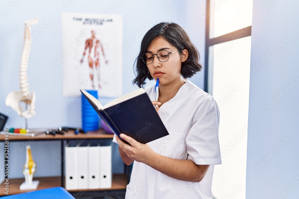 Young latin woman wearing physiotherapist uniform reading book at physiotherapy clinic Young latin woman wearing physiotherapist uniform reading book at physiotherapy clinic