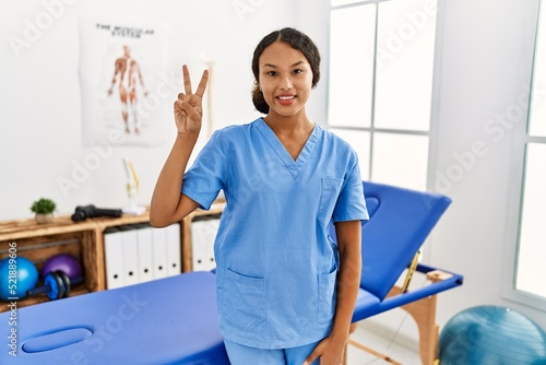 Beautiful hispanic physiotherapist woman at pain recovery clinic showing and pointing up with fingers number two while smiling confident and happy.