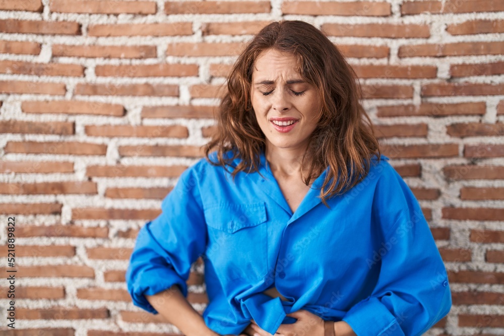 Beautiful brunette woman standing over bricks wall with hand on stomach because indigestion, painful illness feeling unwell. ache concept.