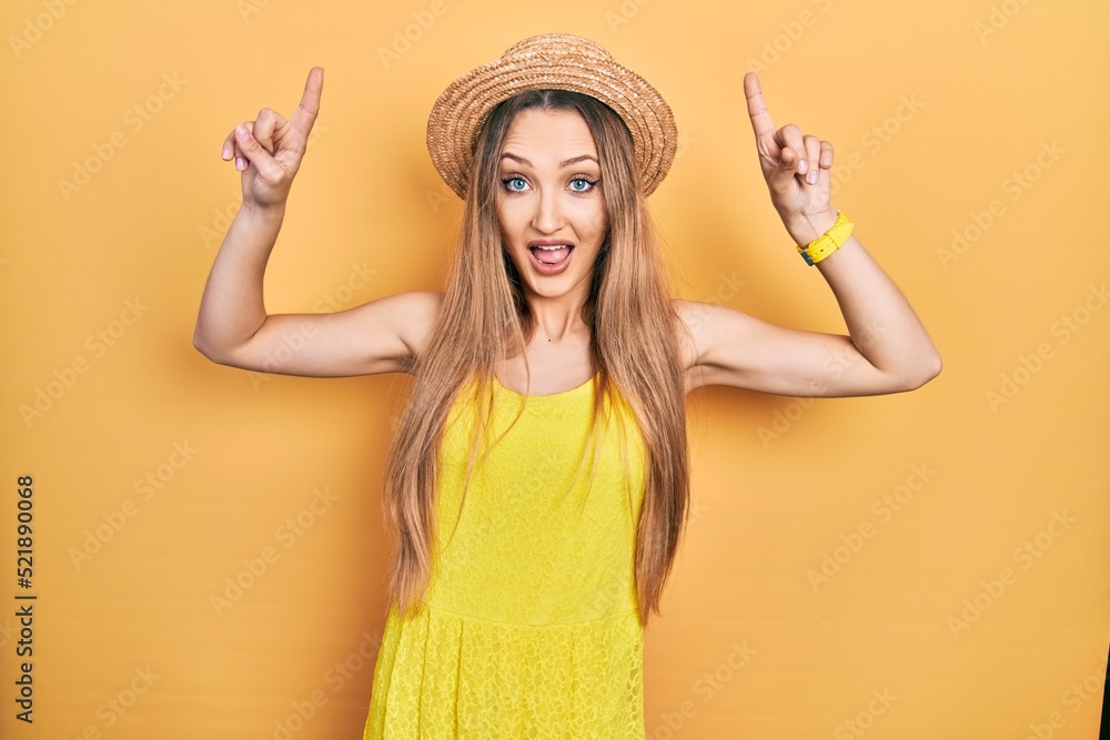Young blonde girl wearing summer hat smiling amazed and surprised and pointing up with fingers and raised arms. Young blonde girl wearing summer hat smiling amazed and surprised and pointing up with fingers and raised arms.