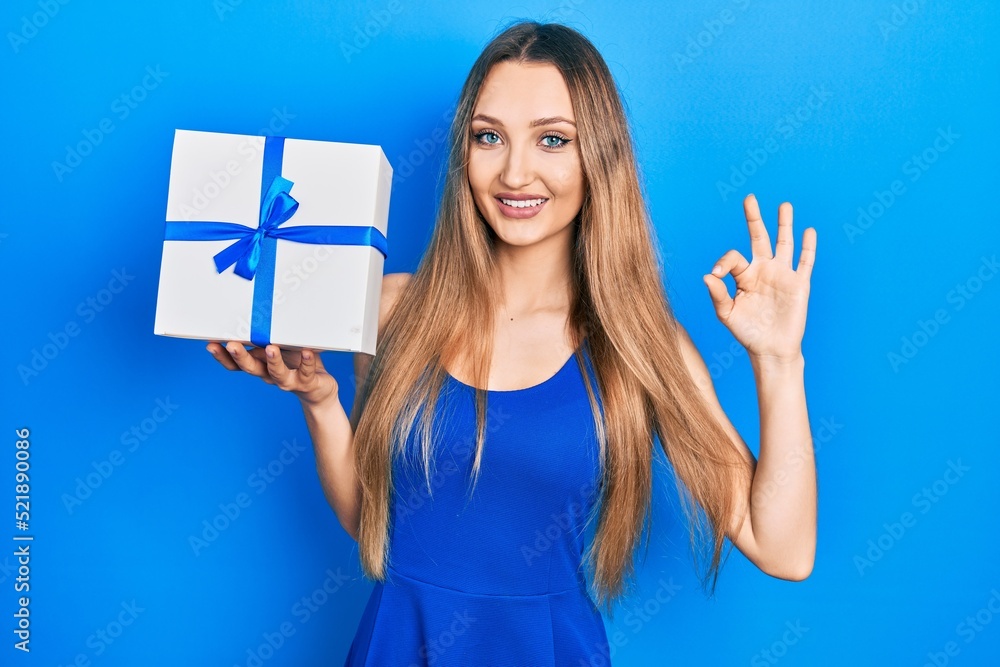 Young blonde girl holding gift doing ok sign with fingers, smiling friendly gesturing excellent symbol Young blonde girl holding gift doing ok sign with fingers, smiling friendly gesturing excellent symbol