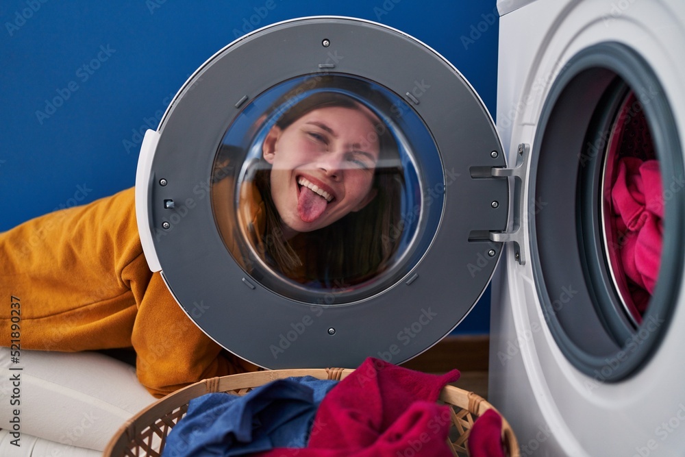 Young brunette woman looking through the washing machine window sticking tongue out happy with funny expression. Young brunette woman looking through the washing machine window sticking tongue out happy with funny expression.