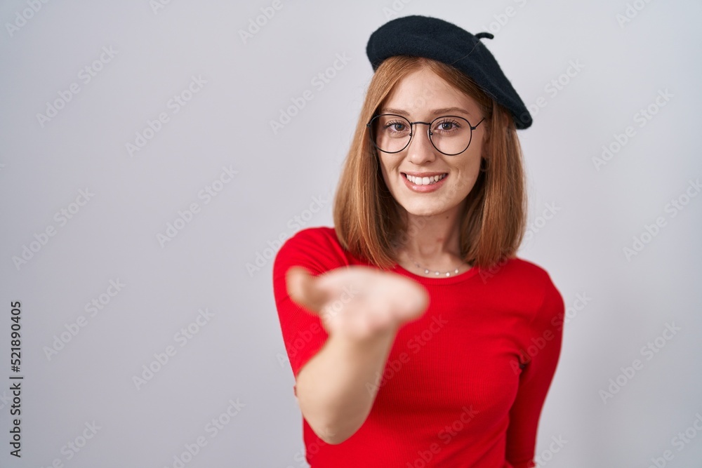 Young redhead woman standing wearing glasses and beret smiling cheerful offering palm hand giving assistance and acceptance. Young redhead woman standing wearing glasses and beret smiling cheerful offering palm hand giving assistance and acceptance.