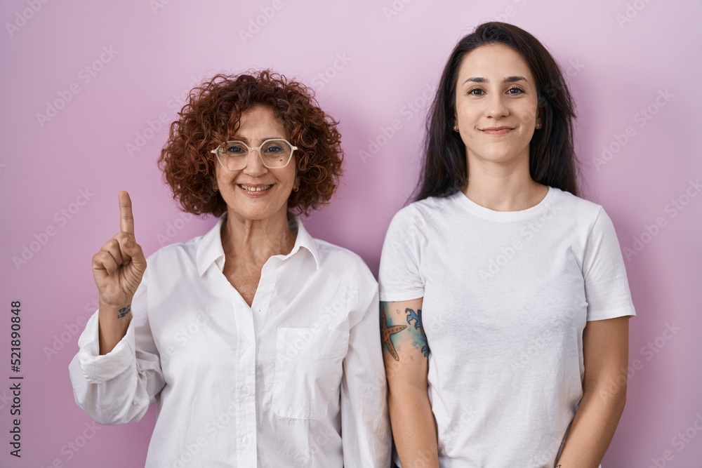 Hispanic mother and daughter wearing casual white t shirt over pink background showing and pointing up with finger number one while smiling confident and happy. Hispanic mother and daughter wearing casual white t shirt over pink background showing and pointing up with finger number one while smiling confident and happy.