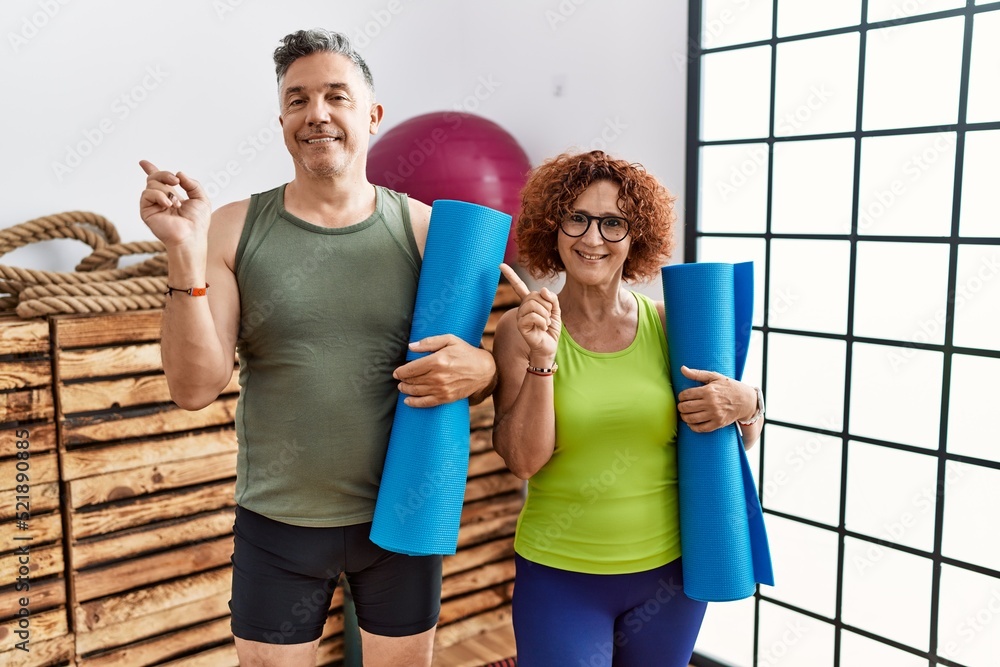 Middle age couple holding yoga mat with a big smile on face, pointing with hand finger to the side looking at the camera. Middle age couple holding yoga mat with a big smile on face, pointing with hand finger to the side looking at the camera.