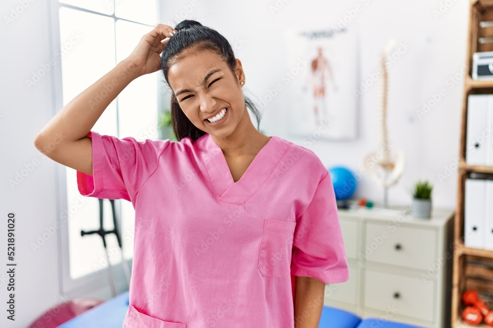 Young hispanic woman working at pain recovery clinic confuse and wonder about question. uncertain with doubt, thinking with hand on head. pensive concept.