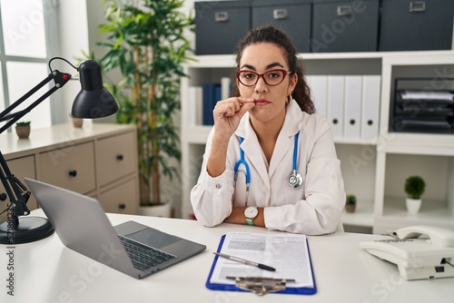 Young hispanic woman wearing doctor uniform and stethoscope mouth and lips shut as zip with fingers. secret and silent, taboo talking