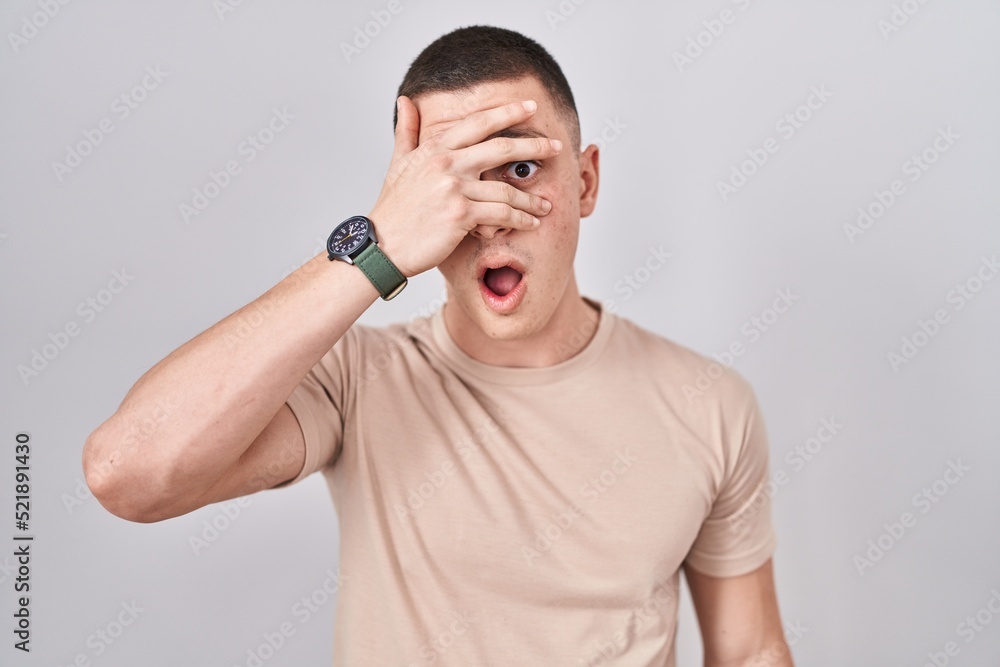 Fototapeta premium Young man standing over isolated background peeking in shock covering face and eyes with hand, looking through fingers with embarrassed expression.