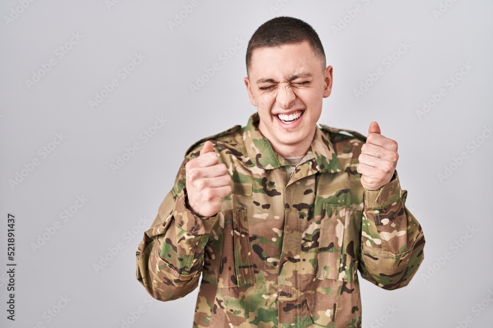 Young man wearing camouflage army uniform excited for success with arms raised and eyes closed celebrating victory smiling. winner concept. Young man wearing camouflage army uniform excited for success with arms raised and eyes closed celebrating victory smiling. winner concept.