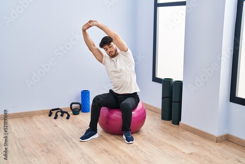 Young hispanic man sitting on fit ball stretching at sport center