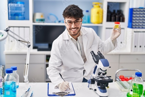 Young hispanic man scientist writing on document holding urine test tube at laboratory