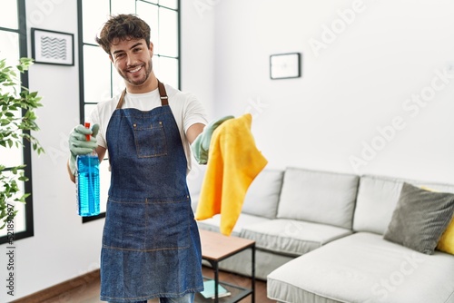 Young hispanic man doing chores holding cleaning sprayer and cloth at home