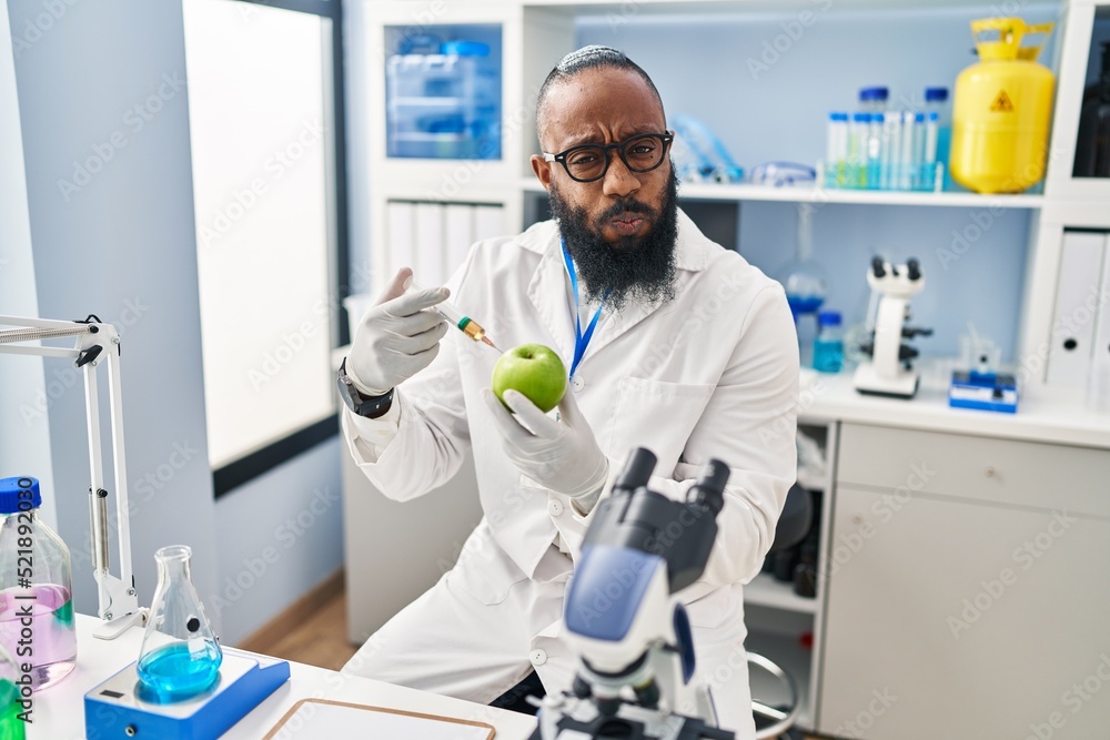 African american man working at scientist laboratory with apple puffing cheeks with funny face. mouth inflated with air, catching air. African american man working at scientist laboratory with apple puffing cheeks with funny face. mouth inflated with air, catching air.