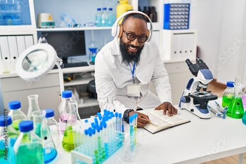 Young african american man wearing scientist uniform reading book listening to music at laboratory