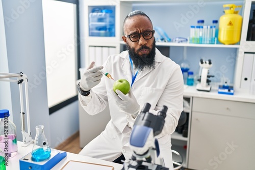African american man working at scientist laboratory with apple puffing cheeks with funny face. mouth inflated with air  catching air.