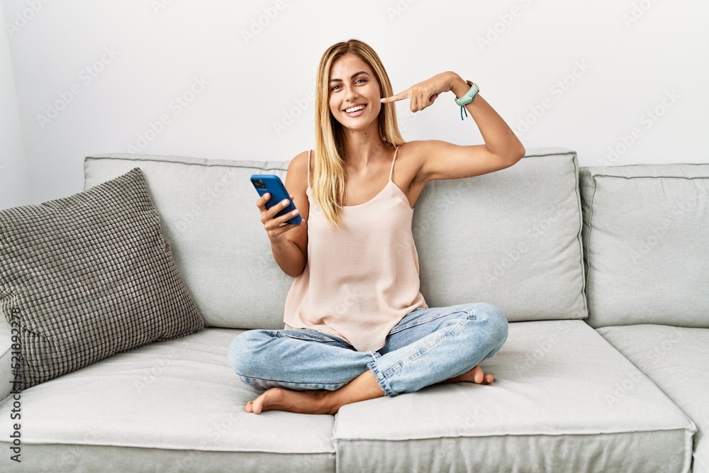 Blonde beautiful young woman sitting on the sofa at home using smartphone smiling cheerful showing and pointing with fingers teeth and mouth. dental health concept.