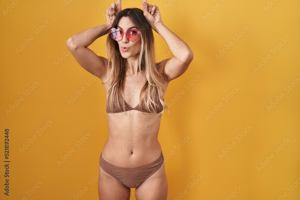 Young hispanic woman wearing bikini over yellow background doing funny gesture with finger over head as bull horns Young hispanic woman wearing bikini over yellow background doing funny gesture with finger over head as bull horns