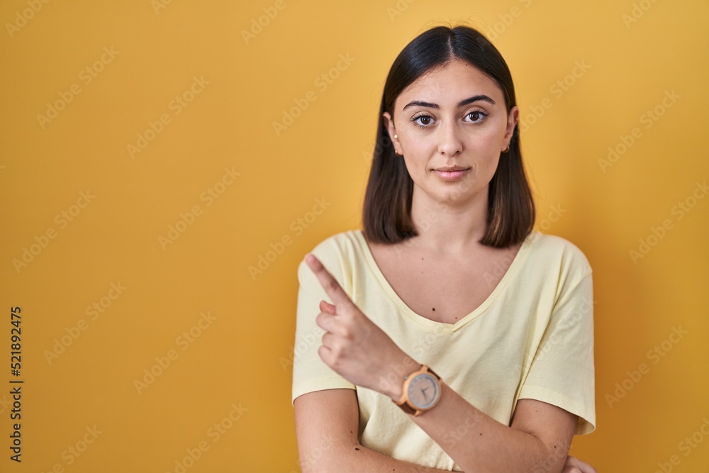 Obraz premium Hispanic girl wearing casual t shirt over yellow background pointing with hand finger to the side showing advertisement, serious and calm face