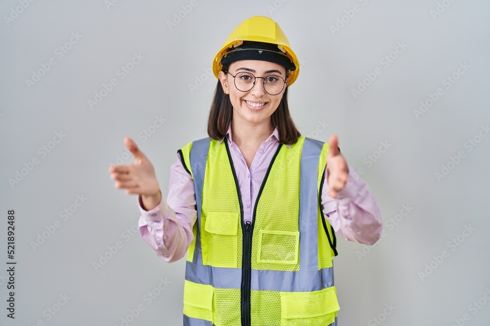 Hispanic girl wearing builder uniform and hardhat looking at the camera smiling with open arms for hug. cheerful expression embracing happiness. Hispanic girl wearing builder uniform and hardhat looking at the camera smiling with open arms for hug. cheerful expression embracing happiness.